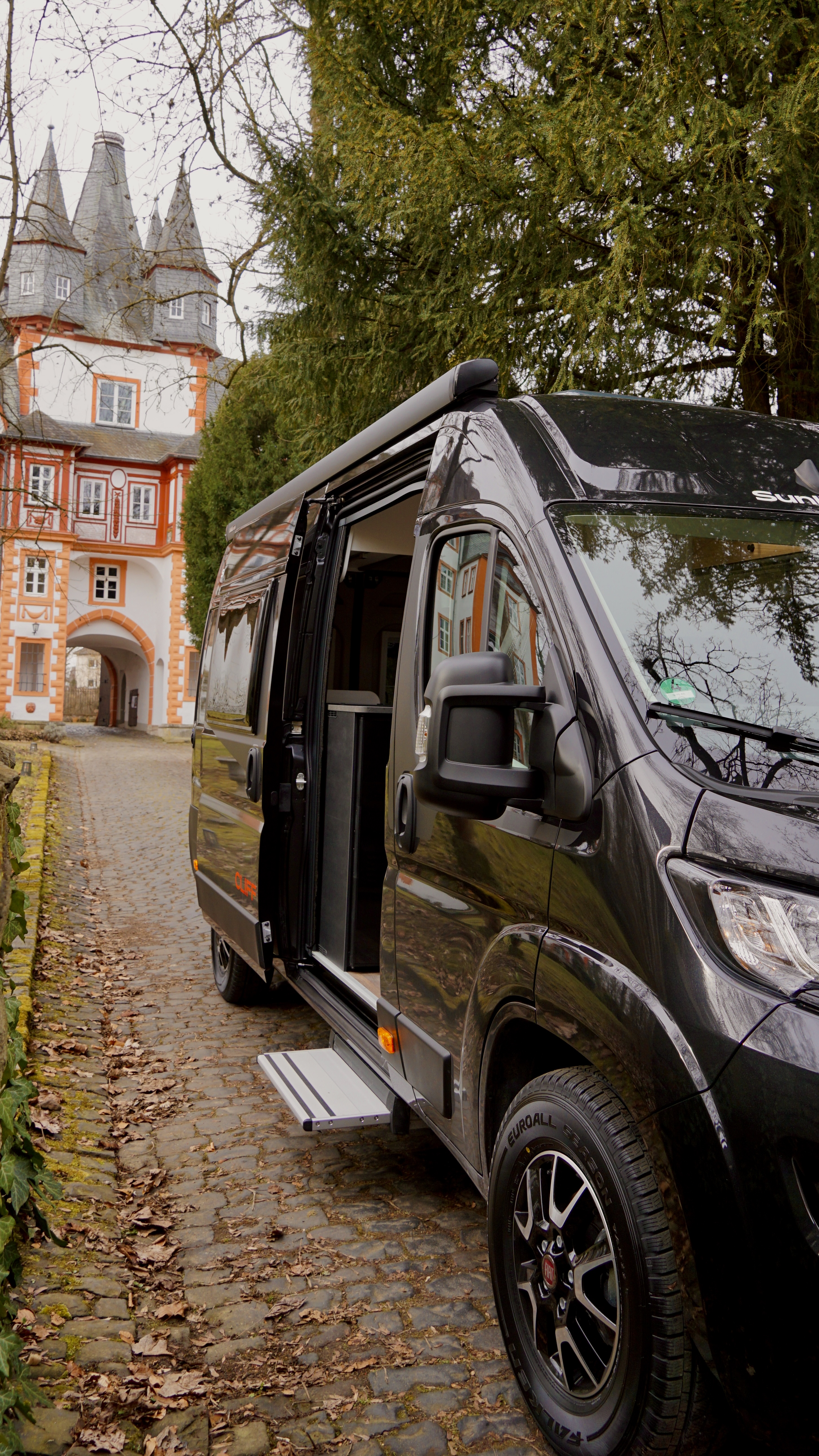 Schwarzer Campervan von Fiat in der Natur vor einem Schloss mit offener Schiebetür
